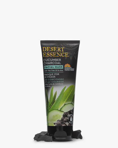 Desert Essence Cucumber Charcoal Face Mask tube standing upright against white background with fresh cucumber slices, green tea leaves, and charcoal pieces scattered at base. Black packaging features gold lettering and green botanical imagery.