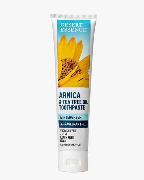 Desert Essence Arnica & Tea Tree Oil Carrageenan Free Toothpaste tube featuring a bright yellow sunflower against a blue sky background. White packaging with blue text highlights natural ingredients and fluoride-free formula in wintergreen flavor.