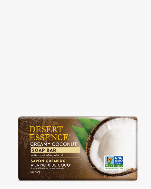 Creamy Coconut Soap Bar with fresh coconut halves and green leaves on brown packaging, featuring Desert Essence branding and Non-GMO verified label, showcasing natural skincare product made with sustainable palm oil.