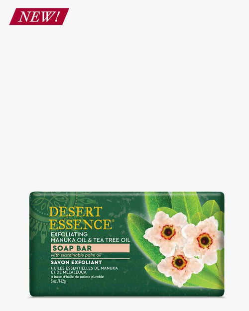 Desert Essence Exfoliating Manuka Oil & Tea Tree Oil Soap Bar shown on dark green packaging with pink Manuka flowers and green leaves. Product features natural ingredients for skin cleansing and exfoliation with sustainable palm oil base. New product label visible.